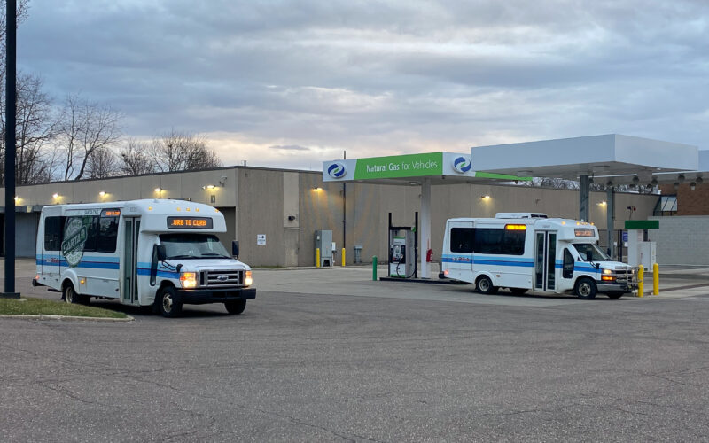 Two white SARTA paratransit buses with blue stripes are parked at a fueling station with a green and white 'Clean Energy' canopy. The SARTA Garage is sitting in the background.