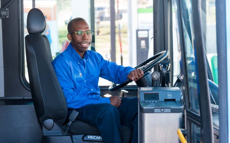 SARTA Driver on a Fixed Route Bus- A male SARTA bus driver wearing a blue uniform sits in the driver’s seat, smiling while holding the steering wheel. The interior of the bus and part of the dashboard are visible, with a fueling station seen in the background through the windshield.